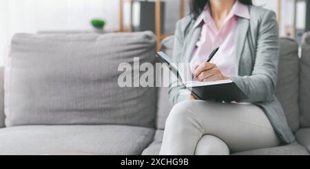 Frau, die in Notizbuch schreibt, während sie auf der Couch sitzt, abgeschnitten Stockfoto