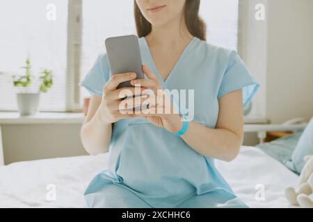 Junge schwangere Patientin, die auf Krankenhausbett und Scrollfutter sitzt Stockfoto
