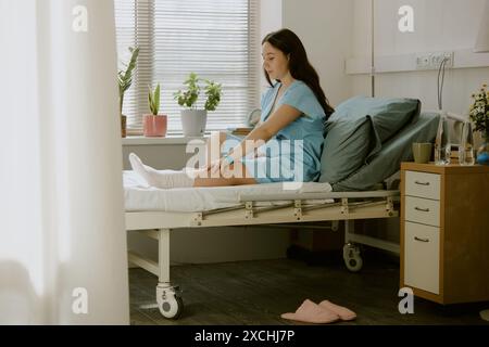 Schwangere Frau, die auf dem Bett sitzt und ihr Bein dehnt, während sie sich auf die Geburt vorbereitet Stockfoto