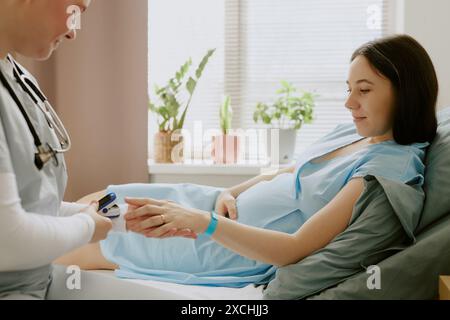 Geburtshelfer untersucht die Sättigung von schwangeren Patienten mittels Pulsoxymeter, während die Patientin auf dem Bett liegt und ihren Bauch hält Stockfoto