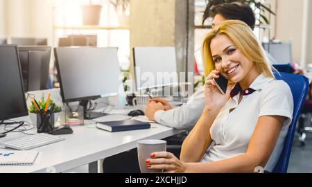 Frau, die am Schreibtisch sitzt und am Handy spricht, Kaffee trinkt Stockfoto