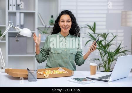 Lächelnde Geschäftsfrau, die im Büro eine Mittagspause macht und ein Telefon hält. Genießen Sie eine Mahlzeit an ihrem Schreibtisch mit einem Laptop und Kaffee. Stockfoto