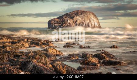 Horizontales Bild von Bass Rock, Schottlands Ostküste, von einem felsigen Ufer aus, an dem die Sonne auf brechenden Wellen scheint. Lange Belichtung Stockfoto