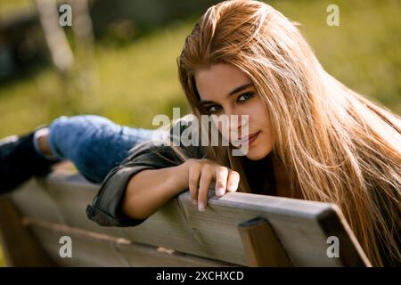 Die junge Frau liegt auf dem Bauch auf einer Parkbank, ihr langes blondes Haar über die Schultern. Sie genießt den Frieden und die Ruhe der Natur Stockfoto