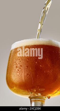 Nahaufnahme eines Bechers gefüllt mit reichhaltigem, bernsteinfarbenem Craft-Bier, das vor grauem Studiohintergrund in Glas gegossen wird. Stockfoto