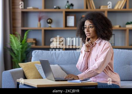 Junge professionelle Frau, die von zu Hause aus mit Laptop arbeitet, tief durchdacht und konzentriert in einem modernen Wohnzimmer. Stockfoto