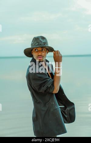 Modernes Porträt eines afroamerikanischen Mannes in schwarzer Jacke und Hut. Junge männliche Model posiert künstlerisch auf natürlichem Hintergrund Stockfoto