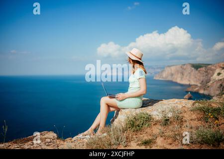 Eine Frau sitzt auf einem Felsen mit Blick auf den Ozean und einem Laptop vor sich. Sie trägt ein grünes Kleid und einen Strohhut. Das Konzept der Entspannung Stockfoto