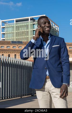 Eleganter afroamerikanischer Manager, der sich im Geschäftsbereich zwischen Firmengebäuden und Büros über ein Smartphone unterhält. Stockfoto