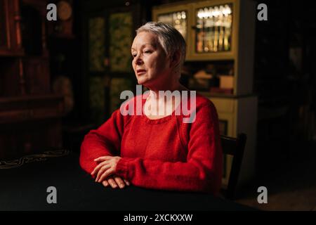 Porträt einer attraktiven, grauhaarigen, erwachsenen Frau in lässigem rotem Pullover, die im Dunkeln Raum mit brennender Kerze am Tisch sitzt Stockfoto