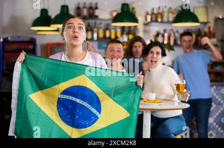 Ein Unternehmen enthusiastischer junger erwachsener Sportfans, die die Flagge Brasiliens schwenken und die Nationalmannschaft mit Bier im Pub unterstützen Stockfoto