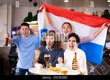 Eine Gruppe von Freunden, die mit der Flagge der Niederlande ein Lieblingsteam anfeuern, während sie sich in der Bar ein Spiel im Fernsehen anschauen Stockfoto