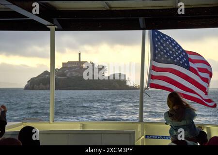 San Francisco, Kalifornien, Vereinigte Staaten von Amerika - 14. Juni 2024: Alcatraz Prison Island at Sunset, aufgenommen von einem Boot mit amerikanischer Flagge *** Gefängnisinsel Alcatraz bei Sonnenuntergang, aufgenommen von einem Boot aus, das eine amerikanische Flagge trägt Stockfoto