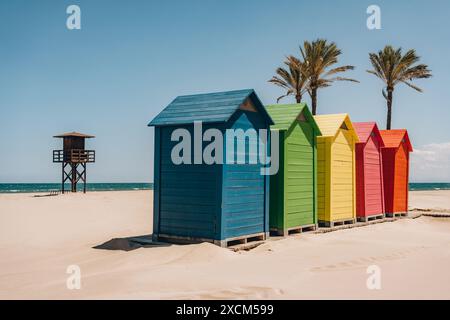 Eine Reihe von farbenfrohen Strandhütten mit Blick auf das Meer, mit Palmen und einem Rettungsschirm-Turm im Hintergrund. Strand Sant Antoni, Cullera, Spanien Stockfoto