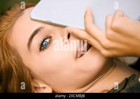 Eine junge Frau mit blonden Haaren und blauen Augen verbirgt ihr Gesicht spielerisch mit einer Tablette und schafft so ein Gefühl von Geheimnissen und Privatsphäre. Sie verkörpert die moderne Ziffer Stockfoto