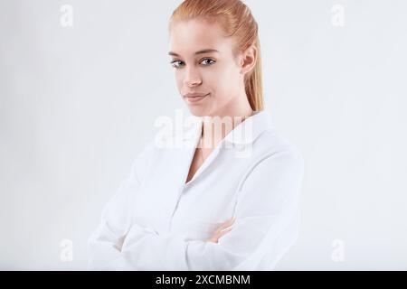 Selbstbewusster Wissenschaftler in Laborkittel lächelnd, Arme gekreuzt, strahlt Expertise in Gesundheitswesen und Wissenschaft vor weißem Hintergrund aus. Junge Erwachsene Rothaarige Show Stockfoto