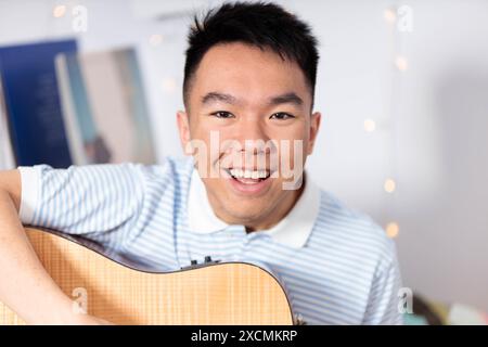 Nahaufnahme eines jungen Teenagers, der fröhlich mit einer Gitarre in seinem gemütlichen Schlafzimmer posiert und direkt mit einem Lächeln auf dem Gesicht in die Kamera blickt Stockfoto