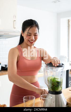 Eine attraktive, schlanke asiatische Frau in Sportbekleidung bereitet ihren grünen Saft mit einem Mixer in der Küche zu und bereitet nach dem Training gesunde Energie-Drinks zu. Reparieren Stockfoto