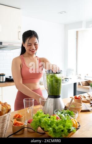 Eine attraktive, schlanke asiatische Frau in Sportbekleidung bereitet ihren grünen Saft mit einem Mixer in der Küche zu und bereitet nach dem Training gesunde Energie-Drinks zu. Reparieren Stockfoto