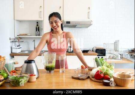 Eine attraktive, schlanke asiatische Frau in Sportbekleidung lächelt die Kamera an und steht in der Küche mit einem Mixer und frischem Gemüse auf der Küche Stockfoto