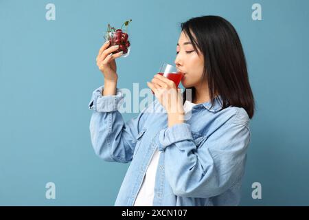 Wunderschöne Asiatin mit Kirschen, die Saft auf blauem Hintergrund trinken Stockfoto