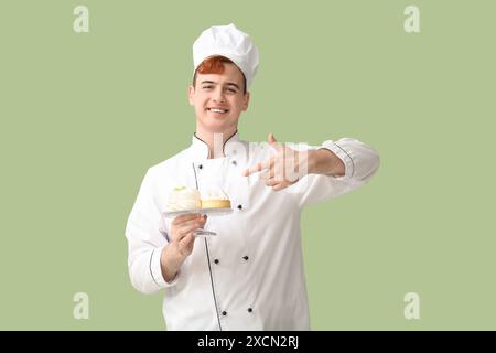 Männliche Konditorin in Uniform, die auf den Stand mit leckeren Desserts auf grünem Hintergrund zeigt Stockfoto