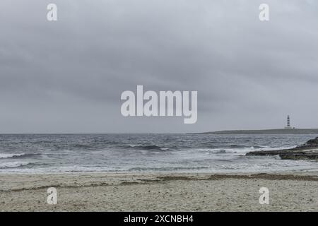 Eine ruhige Szene am Punta Prima Beach auf Menorca, mit Sandstränden, sanften Wellen und einem Fernblick auf einen Leuchtturm unter einem bewölkten Himmel. Stockfoto