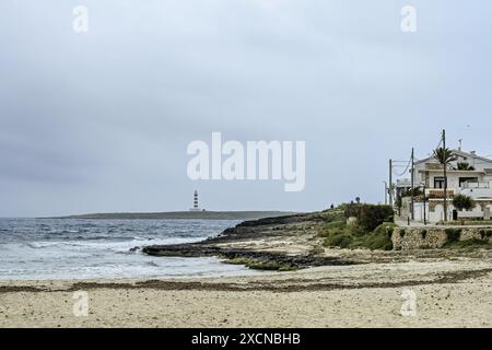 Eine ruhige Szene am Punta Prima Beach auf Menorca, mit Sandstränden, sanften Wellen und einem Fernblick auf einen Leuchtturm unter einem bewölkten Himmel. Stockfoto