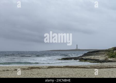 Eine ruhige Szene am Punta Prima Beach auf Menorca, mit Sandstränden, sanften Wellen und einem Fernblick auf einen Leuchtturm unter einem bewölkten Himmel. Stockfoto