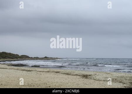 Ein friedlicher Blick auf den Strand Punta Prima auf Menorca, mit einem Sandstrand, sanften Wellen und einem fernen Segelboot unter einem bewölkten Himmel. Stockfoto