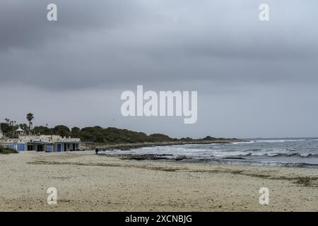 Ein friedlicher Blick auf den Strand Punta Prima auf Menorca, mit einem Sandstrand, sanften Wellen und einem fernen Segelboot unter einem bewölkten Himmel. Stockfoto