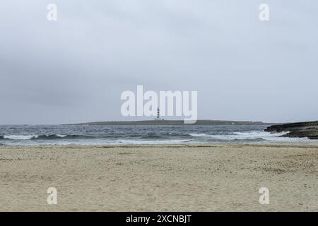Eine ruhige Szene am Punta Prima Beach auf Menorca, mit Sandstränden, sanften Wellen und einem Fernblick auf einen Leuchtturm unter einem bewölkten Himmel. Stockfoto