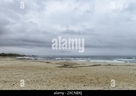 Ein friedlicher Blick auf den Strand Punta Prima auf Menorca, mit einem Sandstrand, sanften Wellen und einem fernen Segelboot unter einem bewölkten Himmel. Stockfoto