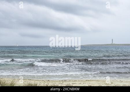 Eine ruhige Szene am Punta Prima Beach auf Menorca, mit Sandstränden, sanften Wellen und einem Fernblick auf einen Leuchtturm unter einem bewölkten Himmel. Stockfoto