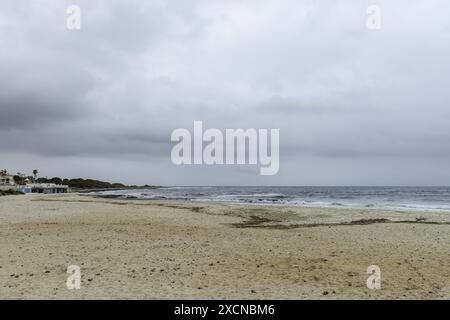 Ein friedlicher Blick auf den Strand Punta Prima auf Menorca, mit einem Sandstrand, sanften Wellen und einem fernen Segelboot unter einem bewölkten Himmel. Stockfoto