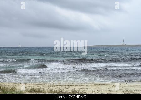 Eine ruhige Szene am Punta Prima Beach auf Menorca, mit Sandstränden, sanften Wellen und einem Fernblick auf einen Leuchtturm unter einem bewölkten Himmel. Stockfoto