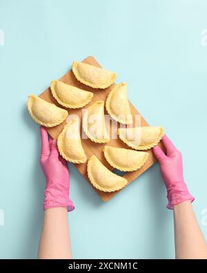 Blick von oben auf Bäckerhände in rosa Gummihandschuhen mit Holzbrett mit rohen Kuchen auf blauem Hintergrund, gefrorene Halbfertigprodukte Stockfoto