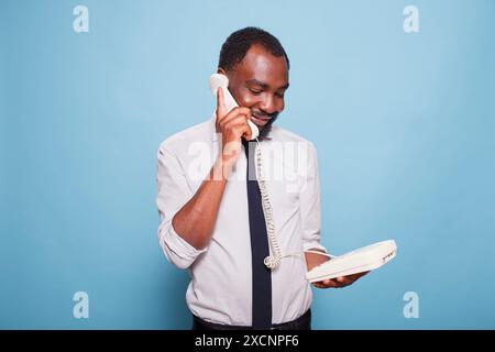 Schwarzer Geschäftsmann, der den Hörer am Ohr hält und über Festnetztelefon spricht, während er den Gesprächspartner am anderen Ende des Anrufs hört. Afroamerikanischer Angestellter in weißem Hemd mit altem Schultelefon. Stockfoto