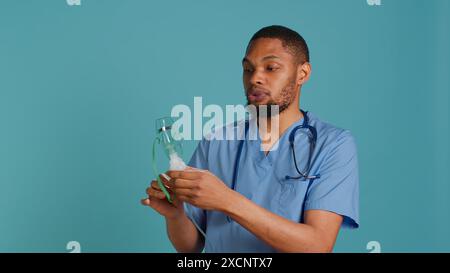 Männliche Krankenschwester, die einen Handvernebler hält, der zur Abgabe von Medikamenten auf Aerosolbasis verwendet wird, um Atembeschwerden zu kontrollieren. Gesundheitsspezialist präsentiert Atemgerät, Studiohintergrund, Kamera A Stockfoto