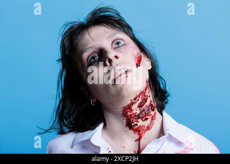 Frau mit blutigem Hals, die vorgibt, tot zu sein, bevor sie sich in Zombie verwandelt und Horrorfilmszene filmt. Schauspielerin, die als untote Leiche auftritt, mit professionellen SFX Make-up Narben, Studiohintergrund Stockfoto