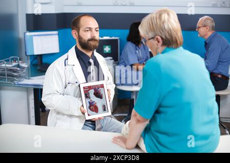 Arzt in weißem Laborkittel erklärt die kardiologische Behandlung, die auf dem Bildschirm des Geräts angezeigt wird. Arzt und älterer Patient analysieren Herzbild auf einem digitalen Tablet. Mann mit Gadget. Stockfoto