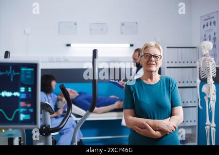 Porträt einer Seniorin mit überkreuzten Armen, die auf körperliches Training wartete. Patientin mit Muskelverletzung mit Blick auf die Kamera bei der Vorbereitung auf die Beinphysiotherapie auf dem Heimtrainer. Stockfoto