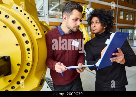 Zwei Ingenieure arbeiten an der Robotik-Programmierung und Automatisierung zusammen und untersuchen Dokumente in der Nähe eines großen gelben Industrieroboters in einer modernen Anlage. Stockfoto