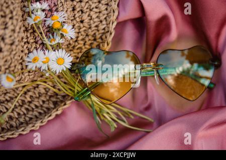 Grüne Sonnenbrille in Herzform, ein Blumenstrauß aus weißen Gänseblümchen und ein Strohhut auf einem rosa seidenen Tuch von oben. Stillleben im Sommer. Sommerurlaub. Stockfoto