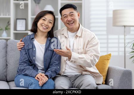 Junge asiatische Paare Mann und Frau, die zu Hause auf der Couch vor der Kamera sitzen und online chatten, stellt der Kerl seine Freundin vor und zeigt mit seiner Hand auf sie. Stockfoto