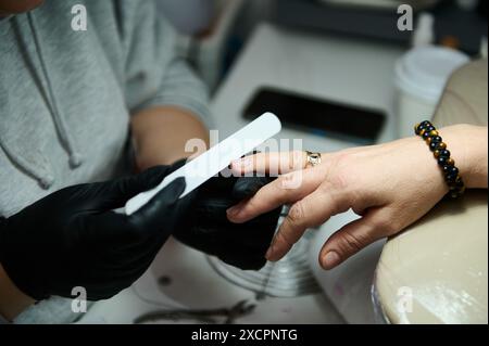 Nahaufnahme eines Manikürs, der Nägel in einem Schönheitssalon feilt. Konzentrieren Sie sich auf Hände, schwarze Handschuhe und Nagelfeile. Professioneller Manikürservice. Stockfoto