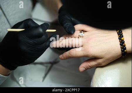 Nahaufnahme eines Manikürers mit schwarzen Handschuhen, der in einem Schönheitssalon an den Nägeln eines Kunden arbeitet. Verbesserung der Nagelschönheit und -Pflege. Stockfoto