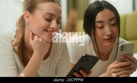 Zwei glückliche lächelnde multirassische Freunde multiethnische Frauen Vielfalt Mädchen weibliche Kaukasierin asiatisch Japanisch Chinesisch entspannen zusammen auf dem Bett mit Handy Stockfoto