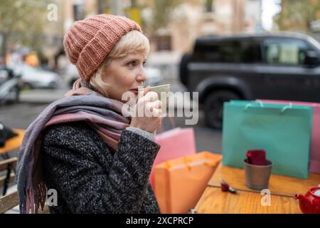 Eine Frau, die draußen eine Kaffeepause genießt, umgeben von Einkaufstaschen. Ideal für Inhalte über Mode, Shopping und Freizeitaktivitäten im Winter. Stockfoto