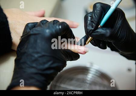 Nahaufnahme einer professionellen Maniküre-Behandlung in einem Schönheitssalon. Der Techniker trägt schwarze Handschuhe und verwendet Werkzeuge, um die Nägel des Kunden zu Formen. Stockfoto